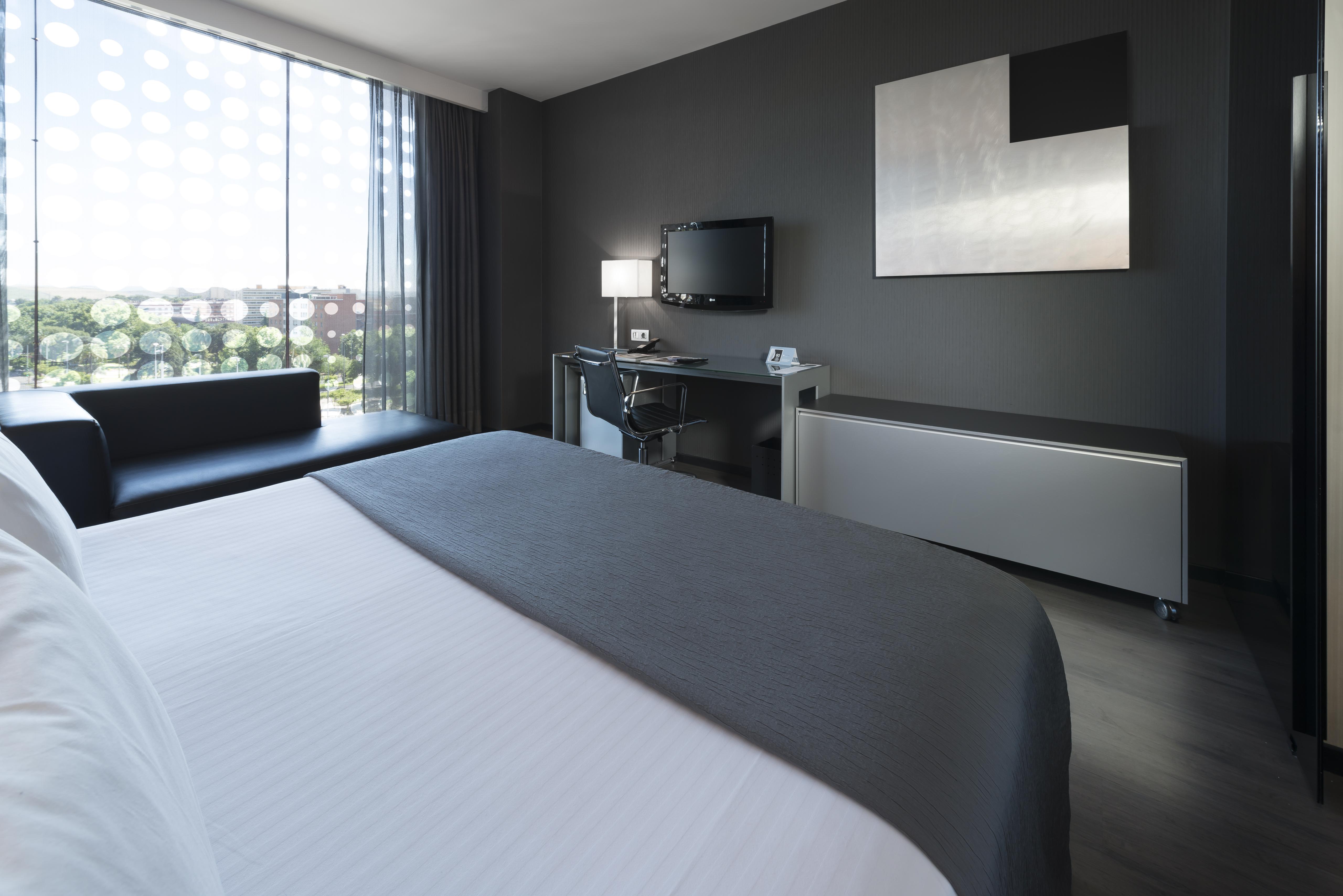 فندق يوروستارز بالاس قرطبة المظهر الخارجي الصورة The photo shows a modern hotel room featuring a sleek design. In the foreground, there is a bed with a white coverlet and gray accents. To the side, a dark sofa is visible. The room has a large window with sheer curtains, allowing natural light to en