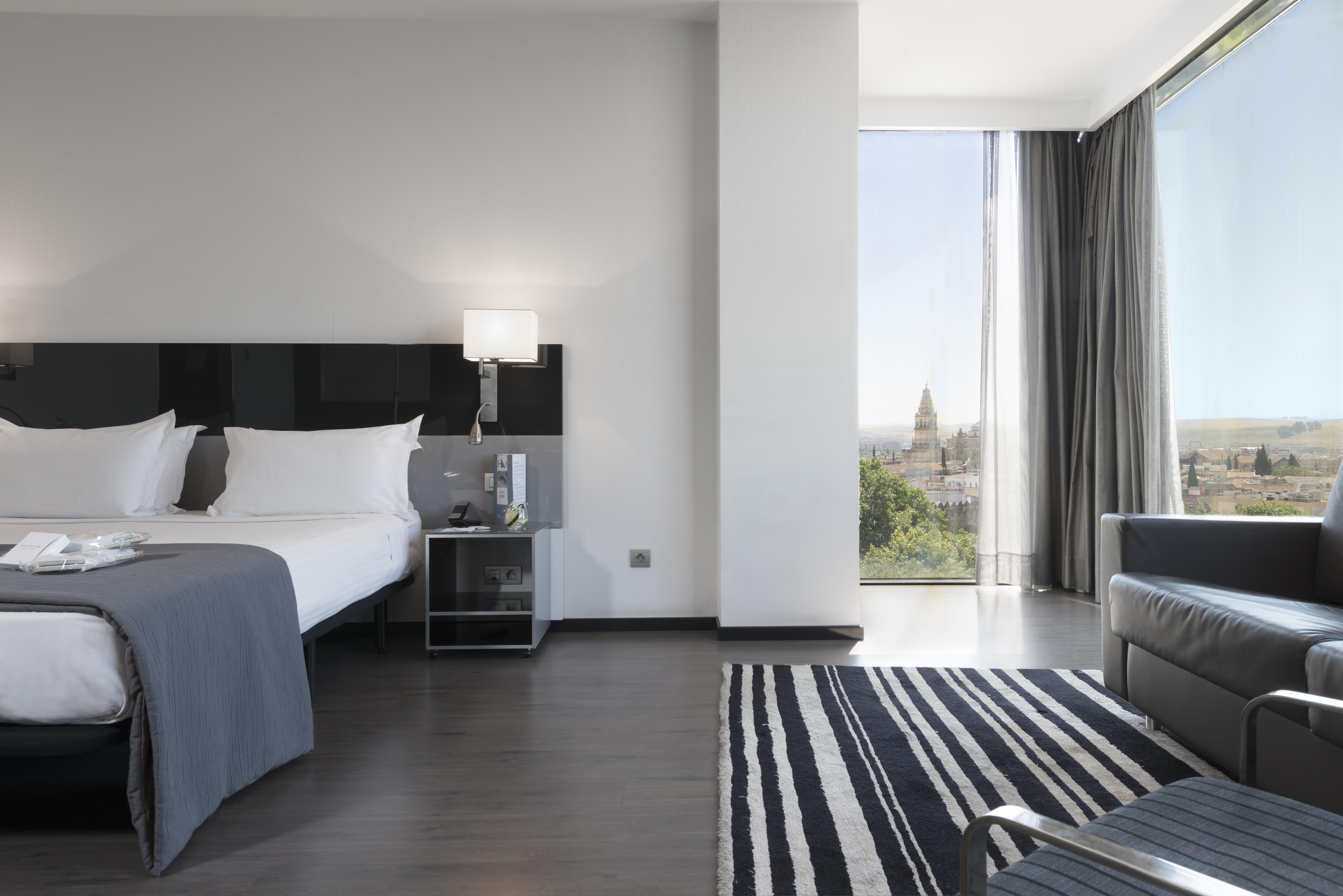 فندق يوروستارز بالاس قرطبة المظهر الخارجي الصورة The photo shows a modern hotel room featuring a large bed with white linens and dark cushions, paired with a sleek nightstand. There is a contemporary chair and a small table nearby, along with a striped area rug on the floor. A large window allows n