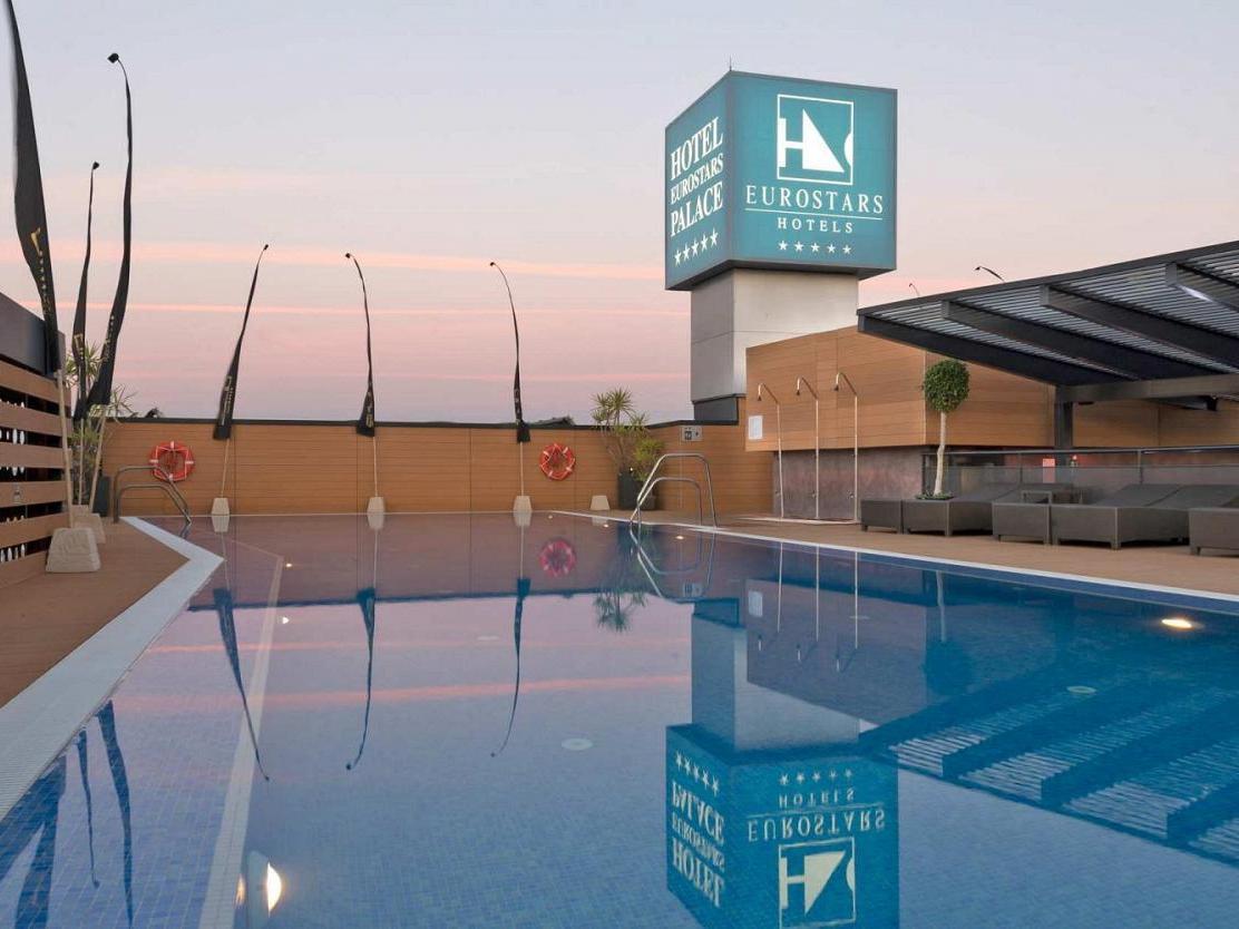 فندق يوروستارز بالاس قرطبة المظهر الخارجي الصورة The image shows a hotel pool area at twilight. The pool is large and reflective, capturing the colors of the sky and surrounding features. In the background, there is a hotel sign displaying the Eurostars brand, with a modern architecture style. The 