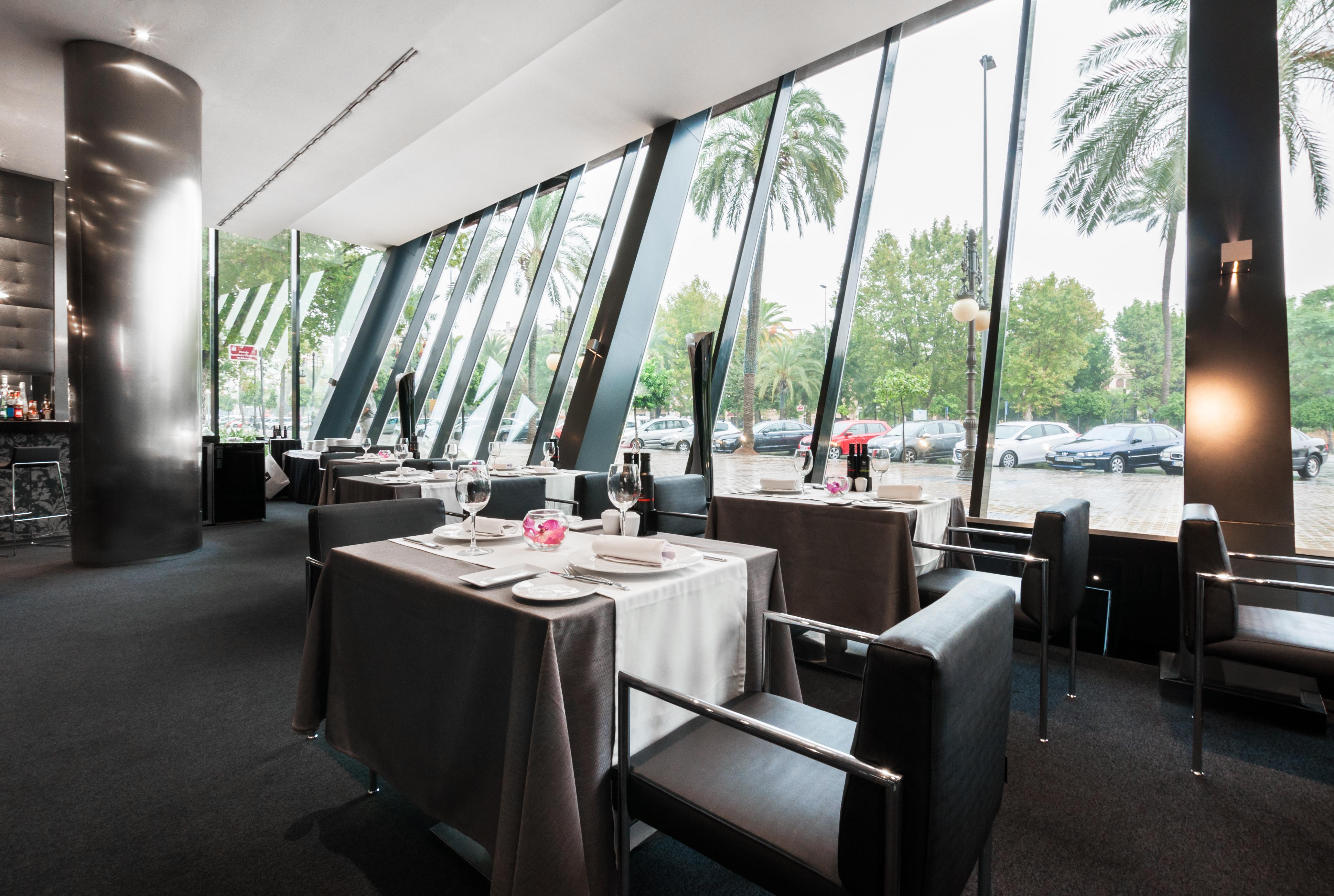 فندق يوروستارز بالاس قرطبة المظهر الخارجي الصورة The photo shows the interior of a modern restaurant featuring large glass windows that provide an expansive view of the outdoors, including palm trees. The dining area is elegantly set up with tables covered in gray tablecloths, decorated with simple