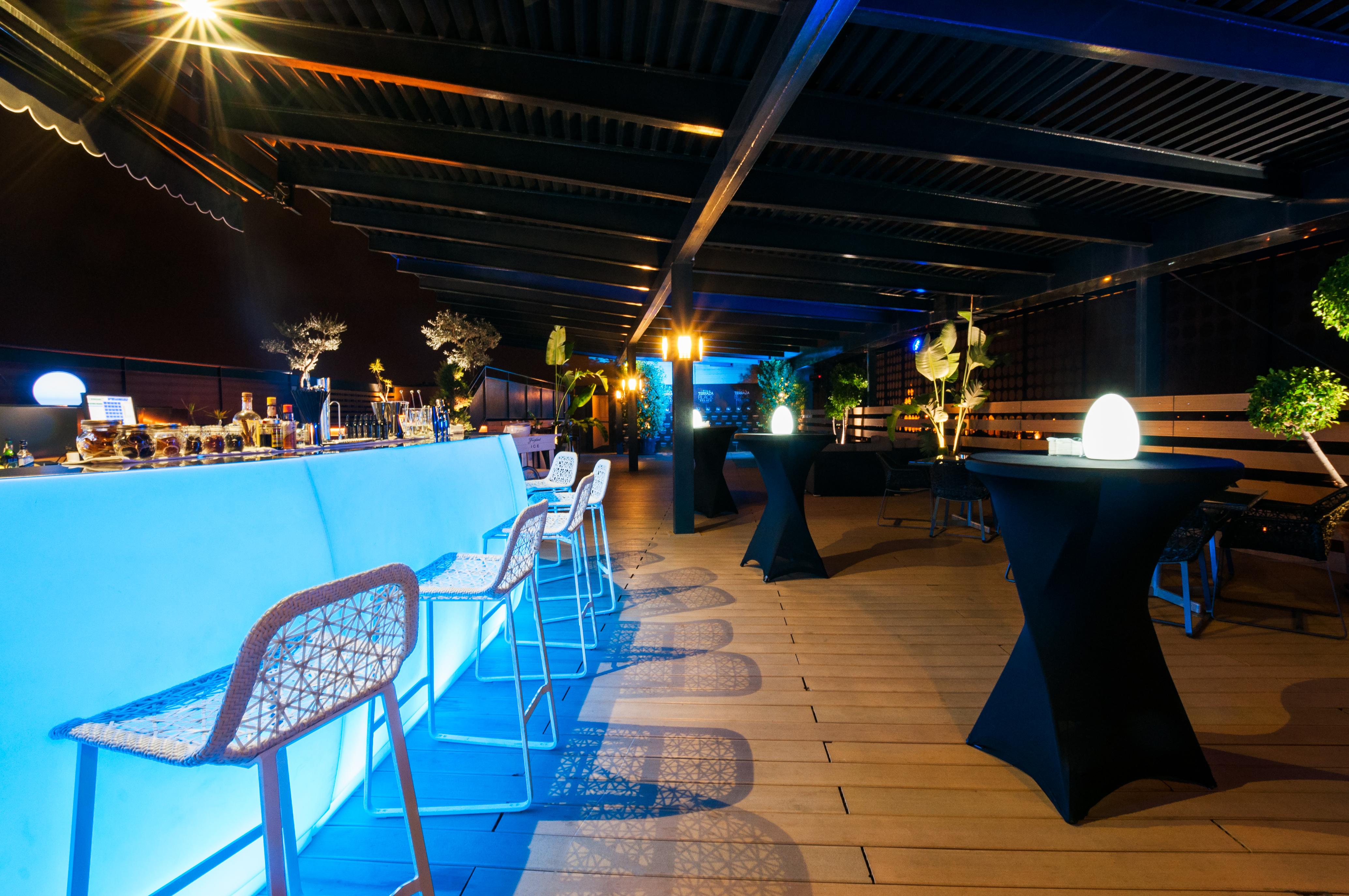 فندق يوروستارز بالاس قرطبة المظهر الخارجي الصورة The photo shows a stylish rooftop or outdoor bar area. There is a long bar counter illuminated with blue lighting, complemented by modern bar stools. The space is decorated with potted plants and elegant round tables covered in black cloth. Soft ligh