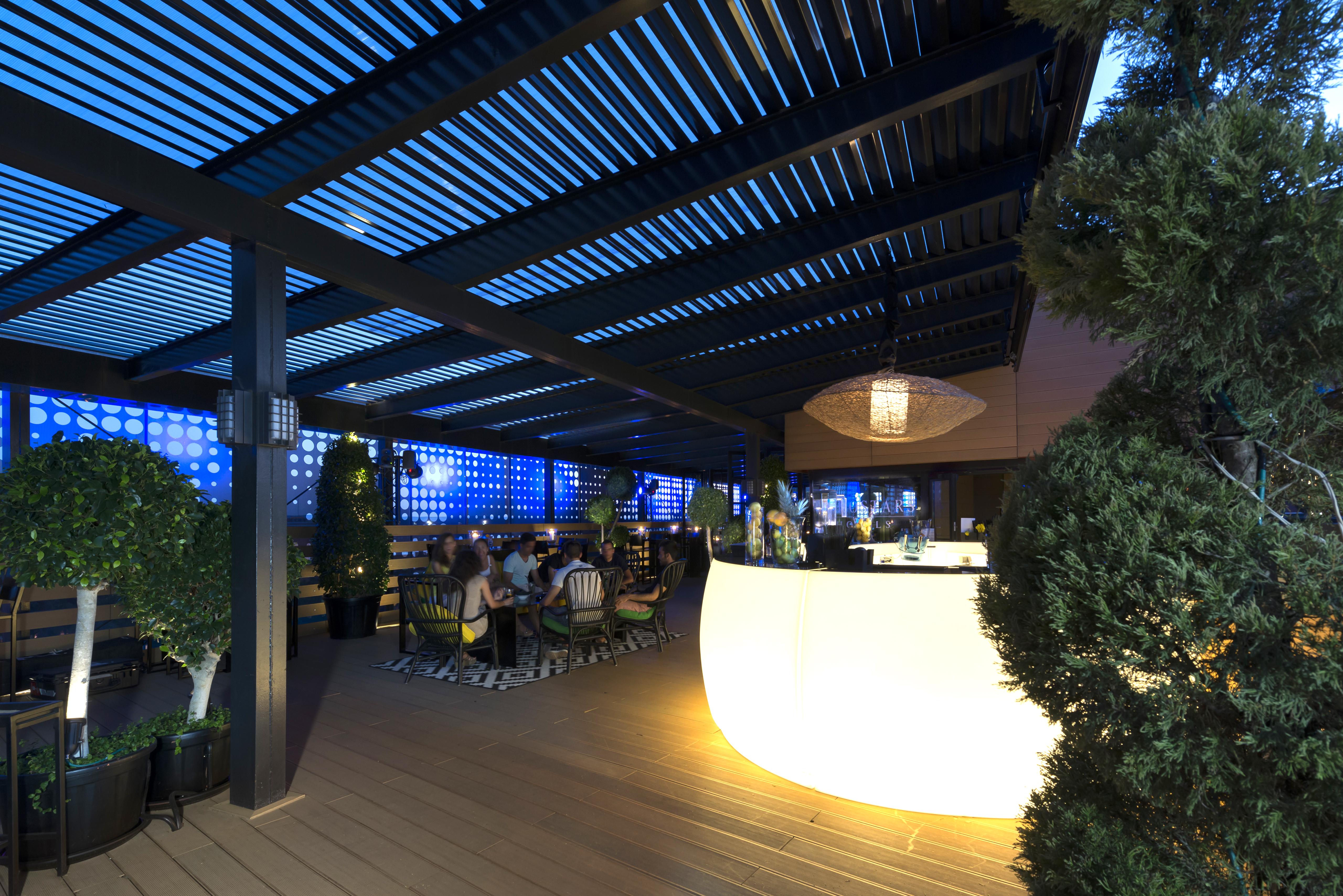 فندق يوروستارز بالاس قرطبة المظهر الخارجي الصورة The image shows a modern outdoor bar or restaurant area. The space features a sleek, illuminated bar that stands out with its curved shape. Patrons can be seen seated around tables, enjoying the ambiance. The overhead structure consists of slatted pa