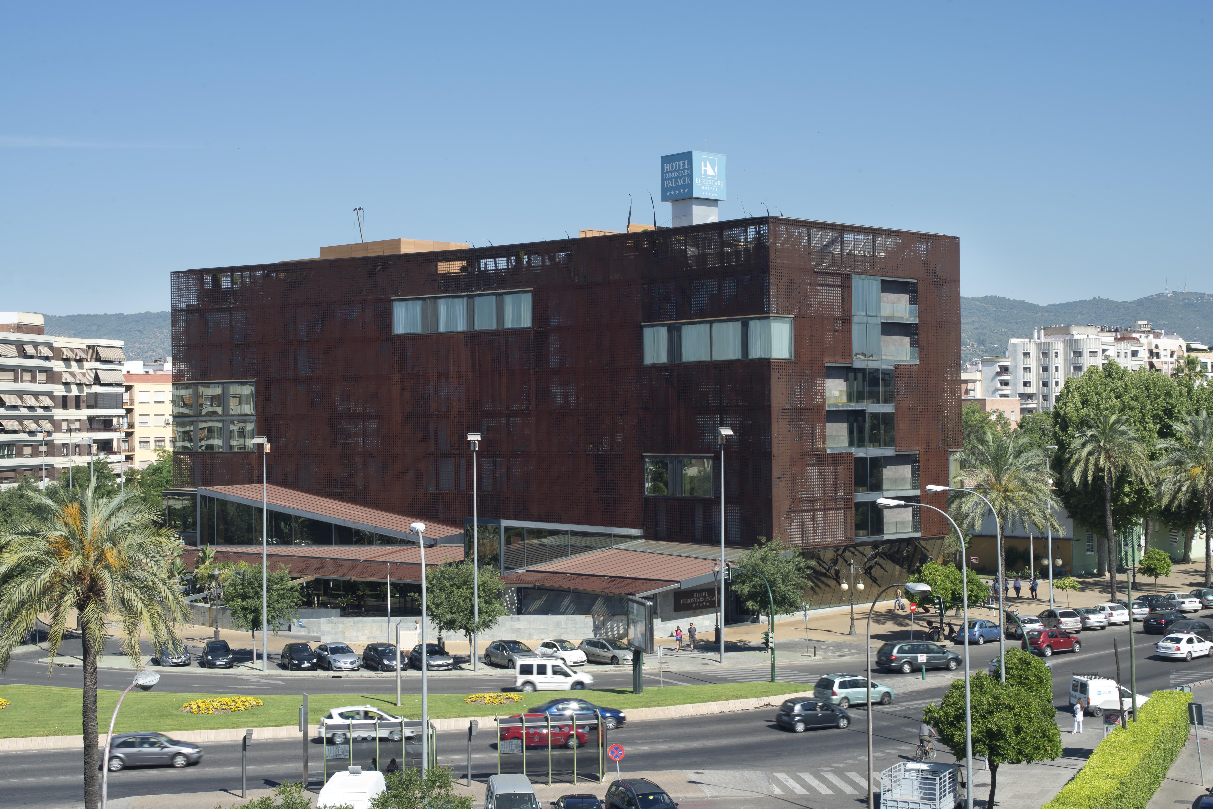 فندق يوروستارز بالاس قرطبة المظهر الخارجي الصورة The photo shows a modern building characterized by a distinctive brown, rust-like exterior, which gives it an industrial appearance. The structure has multiple large windows, allowing for natural light to enter. There are several palm trees surroundi