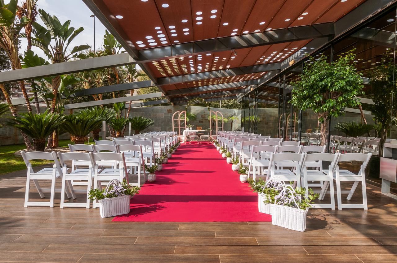 فندق يوروستارز بالاس قرطبة المظهر الخارجي الصورة The image shows an outdoor wedding setup. There are white chairs arranged neatly on either side of a vibrant red carpet that leads to a framed archway. The area is covered by a glass and metal structure, allowing plenty of natural light, while tropic