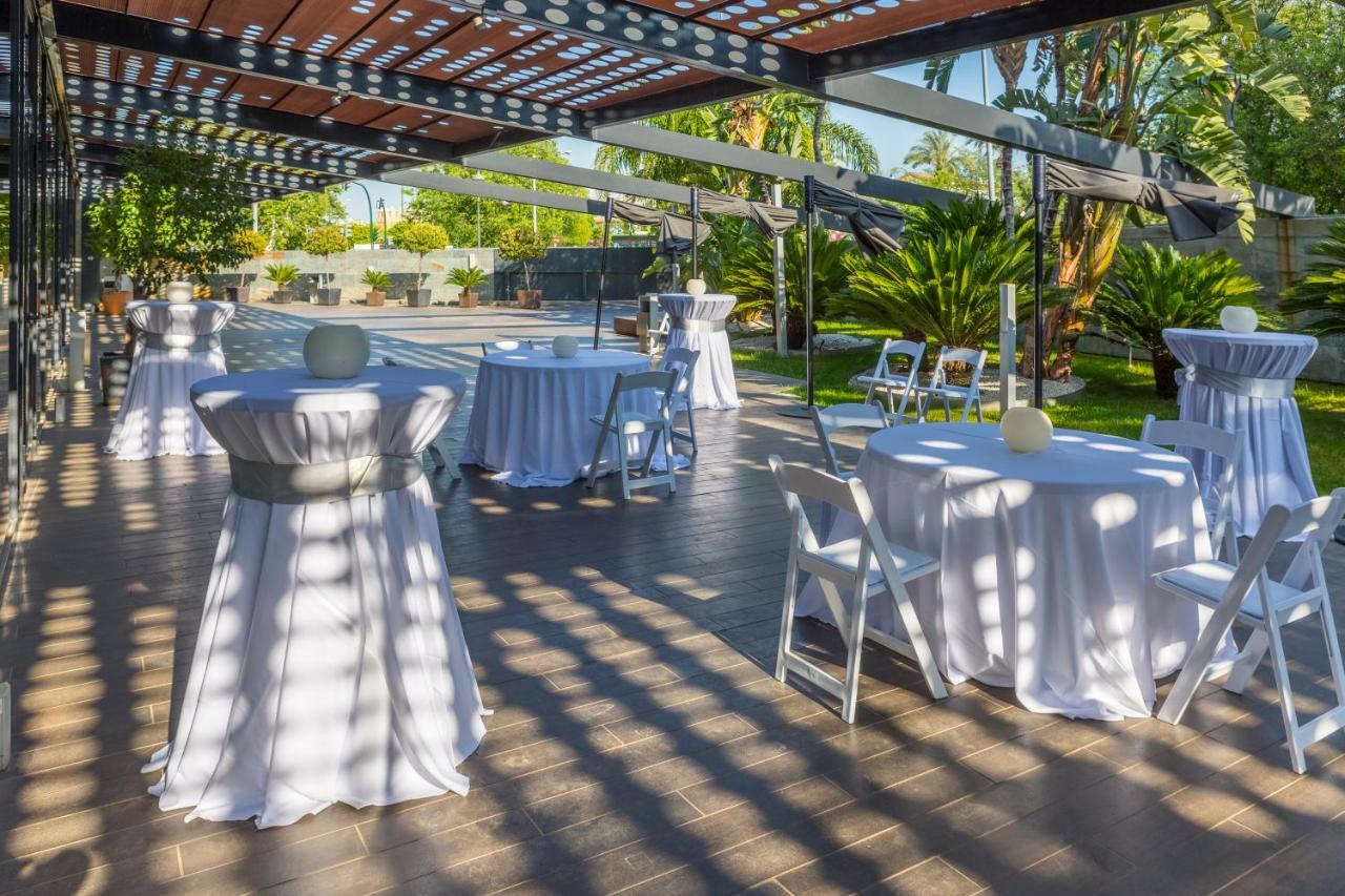 فندق يوروستارز بالاس قرطبة المظهر الخارجي الصورة The photo shows an outdoor event setup. There are several round tables covered with white tablecloths, some adorned with decorative elements like small centerpieces. Chairs are arranged around the tables, and the area is surrounded by greenery, inclu